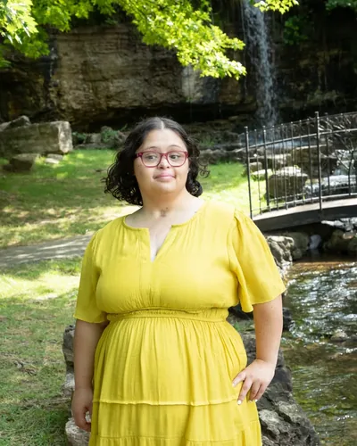 Danielle Robinson standing outdoors in a yellow dress beside a creek and a small waterfall.
